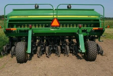 A large, green agricultural machine with exposed wheels and tines sits on a dirt field, displaying a triangular warning sign at the rear.