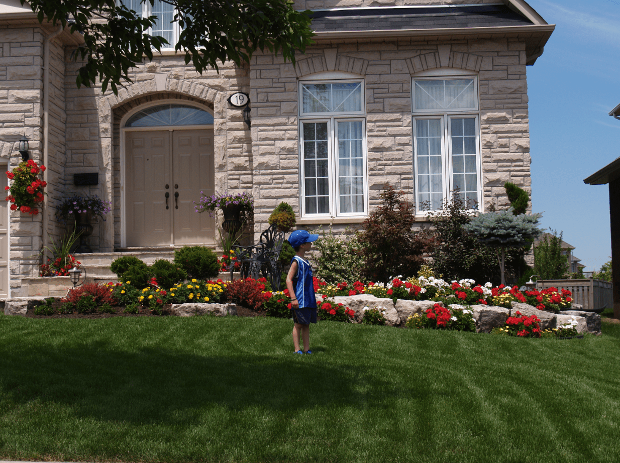 A child stands on a well-manicured lawn in front of a stone house with a flower garden and double doors.