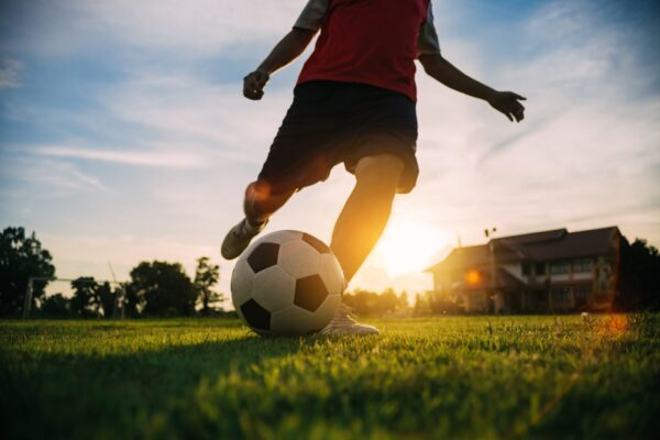 A person playing soccer on a grassy field at sunset, with a large building in the background and trees nearby.