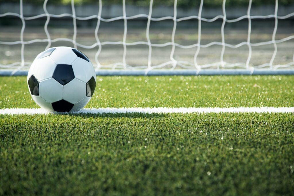 A black and white soccer ball rests on a white line in front of a goal on a green field.