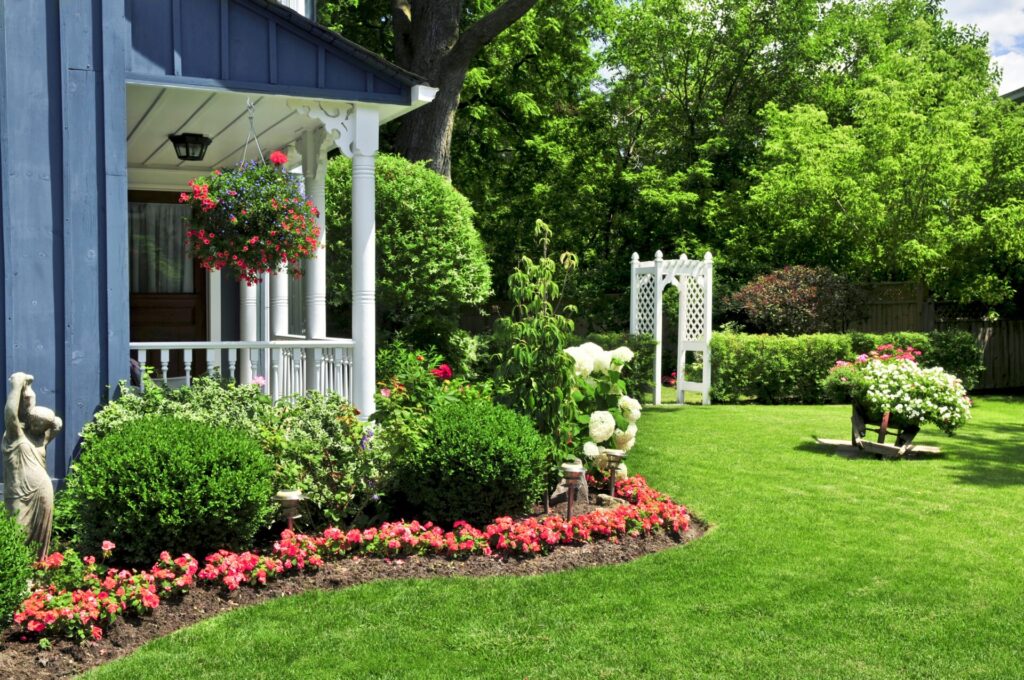 A charming garden with manicured lawns, vibrant flowers, a white trellis, and a blue house with a porch adorned by hanging plants.